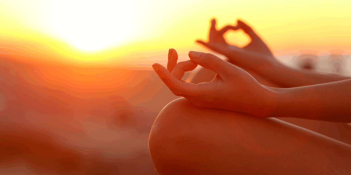 Feminine hands in a meditative pose with an orange sunset in the background.