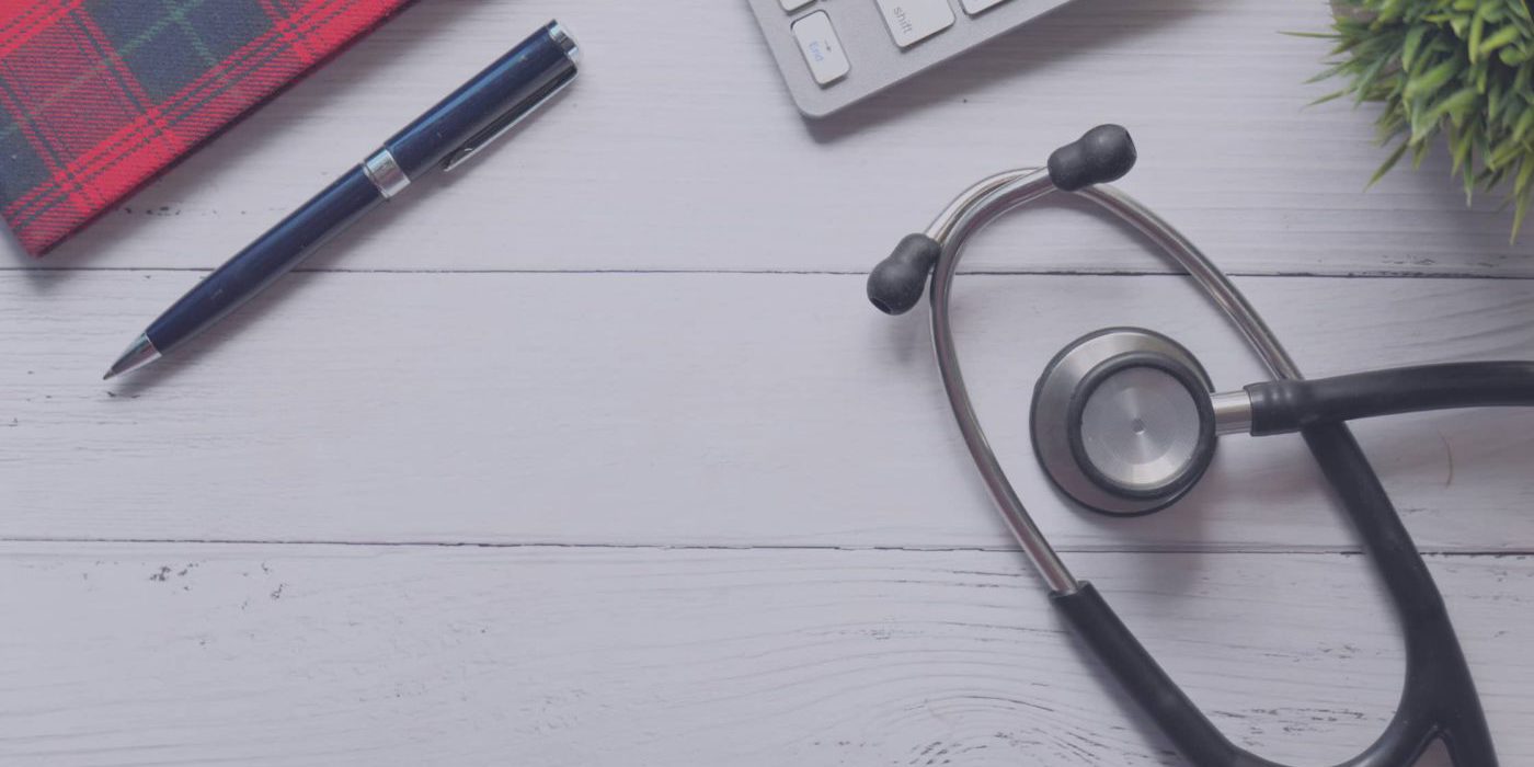 Birds eye view of a clinic desk with notepad, pen, laptop, plant and stethoscope in view.