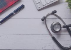 Birds eye view of a clinic desk with notepad, pen, laptop, plant and stethoscope in view.